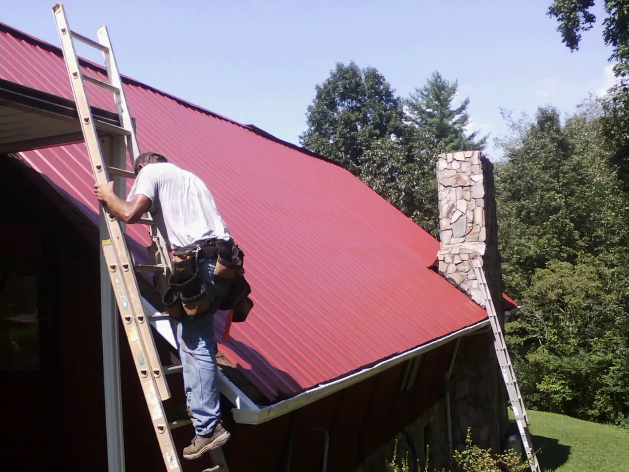 Roofer on a ladder beside a red metal roof and stone chimney — Soffit & Fascia Repair by Roofing Brothers in Alloway